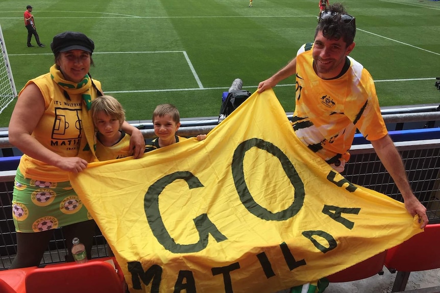 Mithra Cox and her sons and partner in soccer stadium in France with a large flag that says 'GO MATILDAS'.