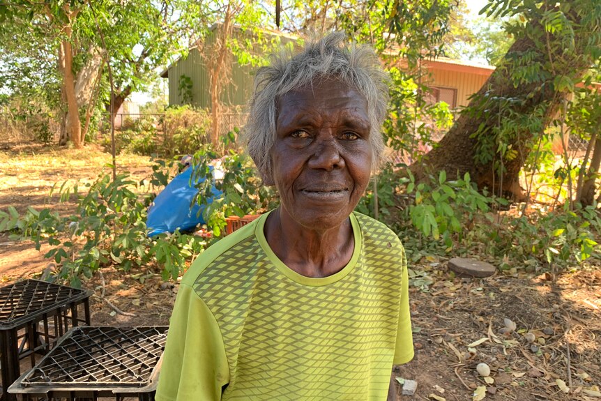 An elderly Indigenous woman standing in a garden, looking at the camera with a smile