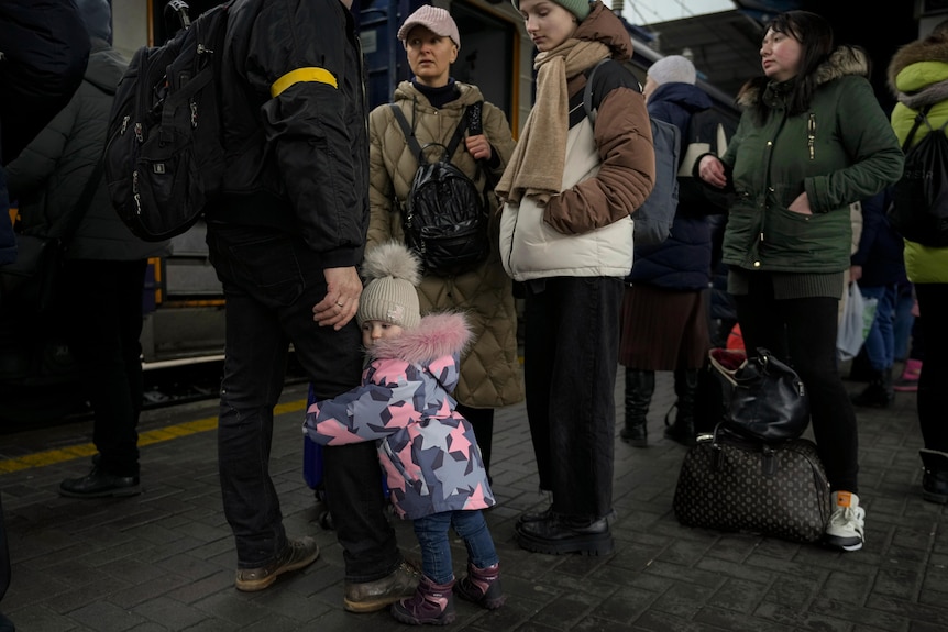 A little girl hugs the leg of a man as adults stand on a train platform.