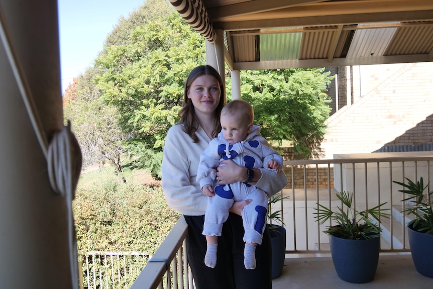 A brunette woman in a white cardigan holding her baby while leaning against a balcony at a home.