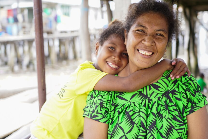 A Papua New Guinean woman grinning while a girl wraps her arms around her neck 