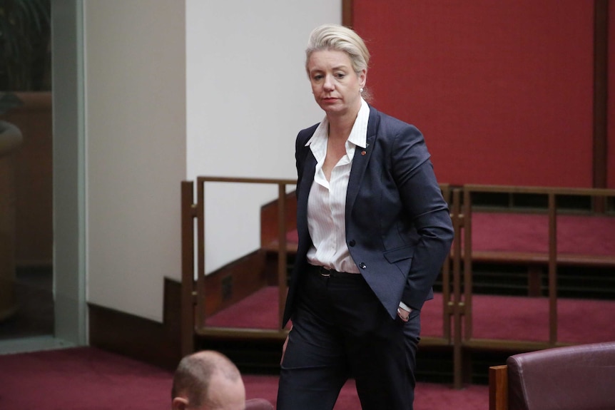 McKenzie is walking, hand in pocket, wearing a blue suit and white shirt with the collar unbuttoned.