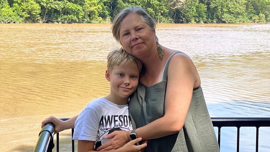 A mother with her arm around her son in front of a river.