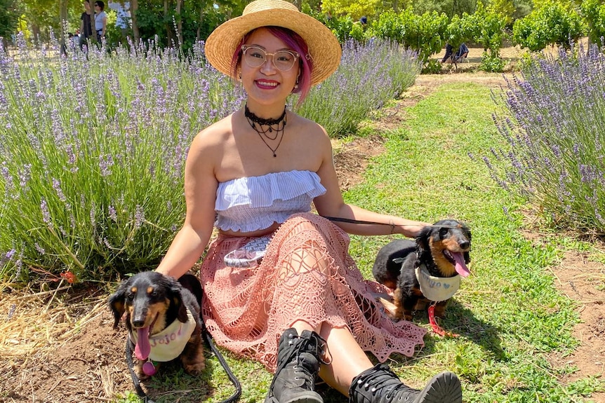 A young woman wearing a floral dress sits at an outdoor cafe table holding two dogs on her lap.