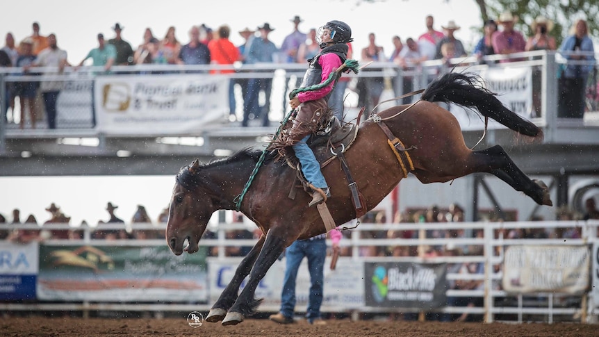 a woman wearing pink is photographed from the side as she rides a bucking horse 