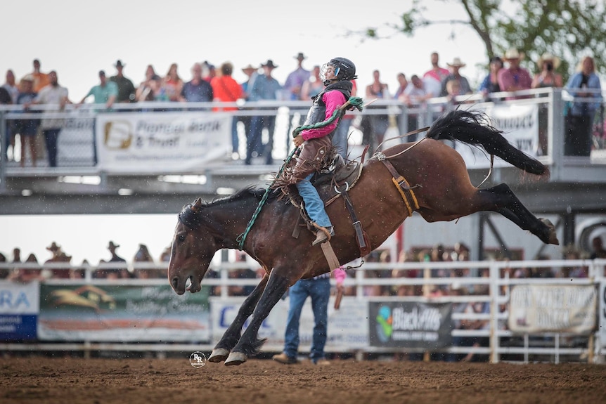 A woman wearing pink is photographed from the side as she rides a bucking horse.