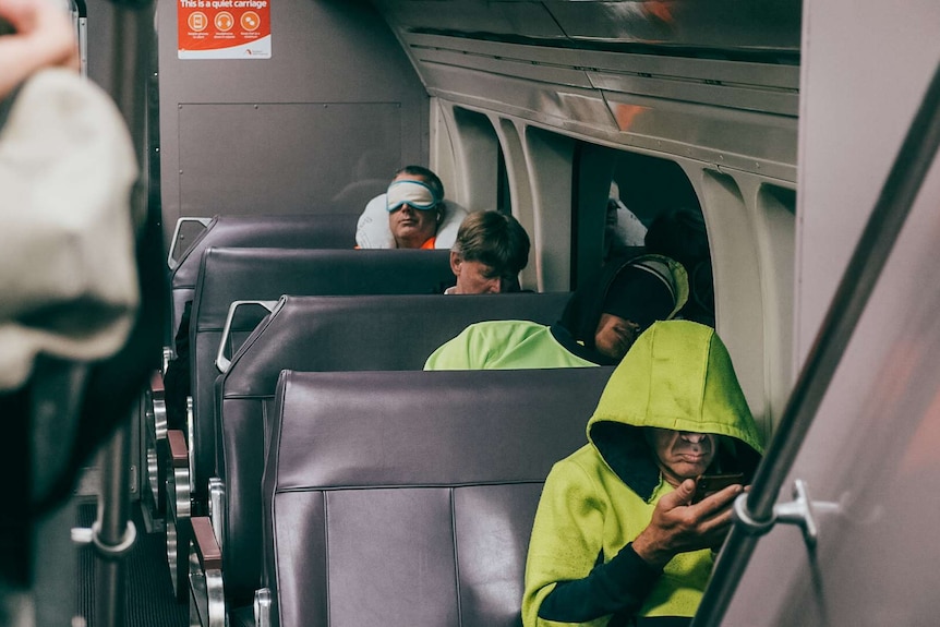 Tradie commuters sleeping and playing on their phones on a dawn train.