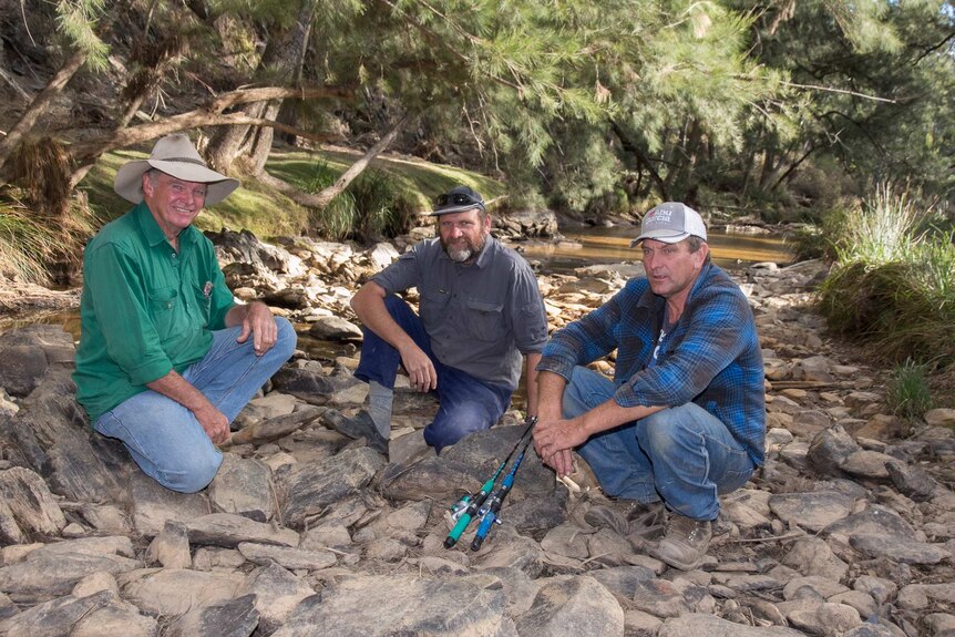 Three men with some fishing rods sit on rocks in front of a river