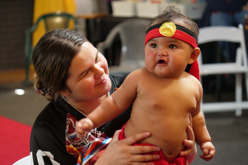 James, with mum holding him, is wearing an Aboriginal flag headband, no shirt, arms out. 