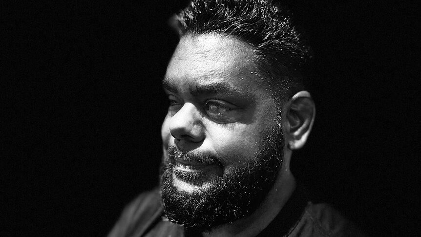 Close up black and white photograph of indigenous man side on with beard, black back ground