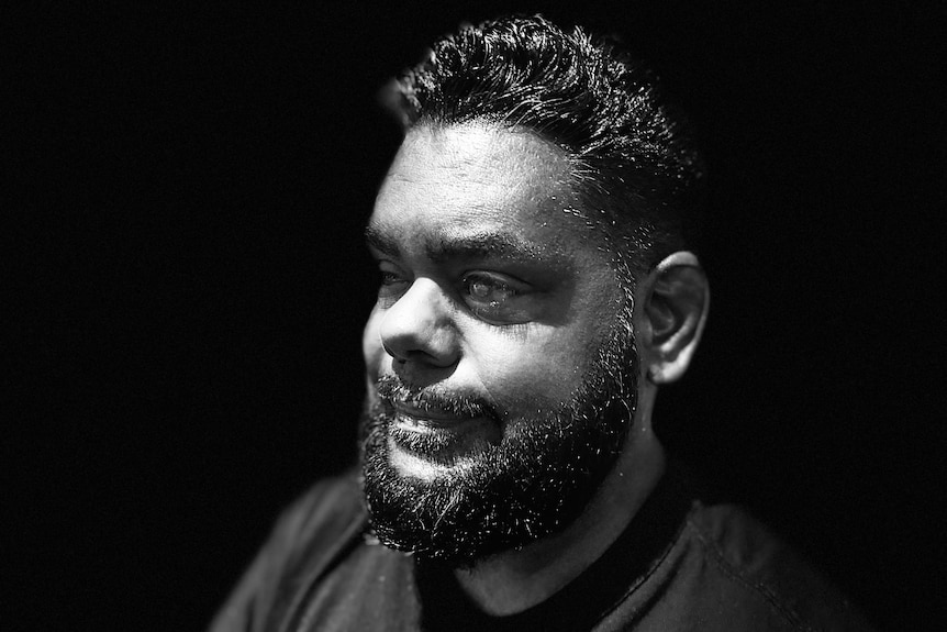 Close up black and white photograph of indigenous man side on with beard, black back ground