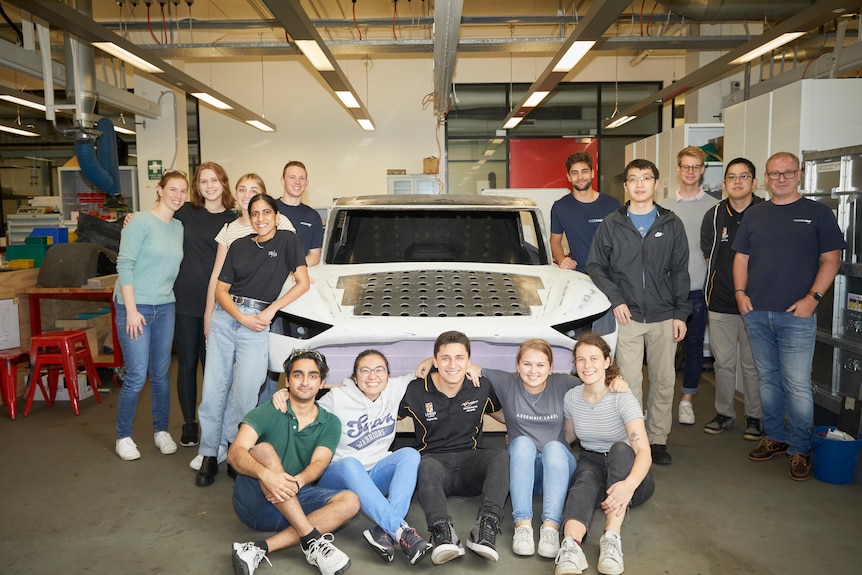 students pose around a white car with solar panels on it inside a workshop.