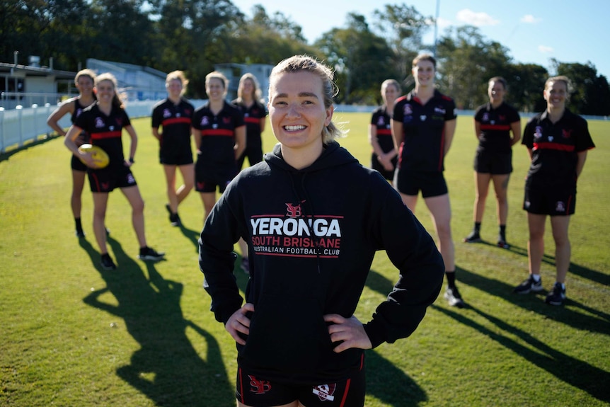 Women wearing black and AFL uniforms stand on an AFL field. One woman stands in the foreground smiling.