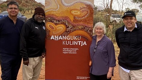 three aboriginal men stand on dirt with a white woman with grey hair with a brown pull-up banner in the middle