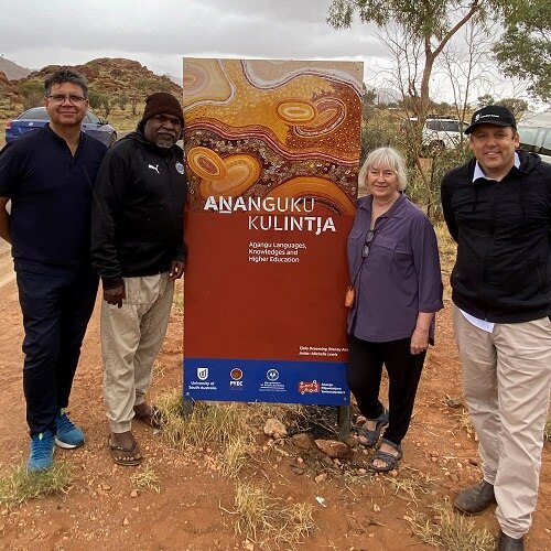 three aboriginal men stand on dirt with a white woman with grey hair with a brown pull-up banner in the middle