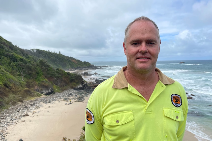 A middle-aged man stands in a yellow rangers shirt, with a rugged, rocky beach behind him.