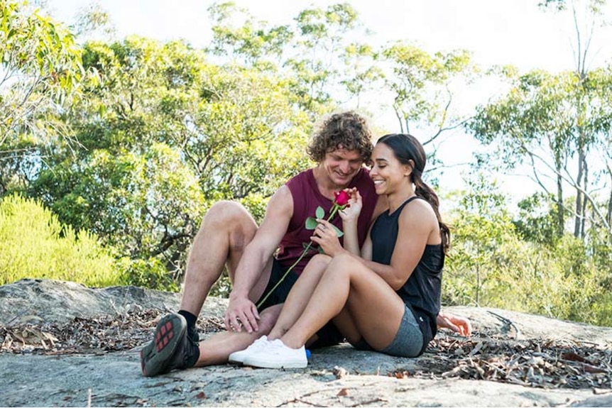 A man with curly hair sits on a big rock in the bush cuddled up to a petite woman with brown hair holding a rose