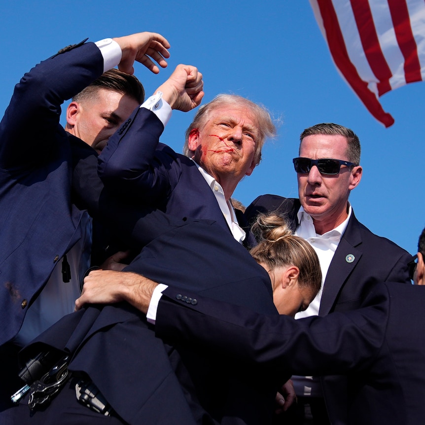 Donald Trump is surrounded by US Secret Service agents on a stage with red around his mouth