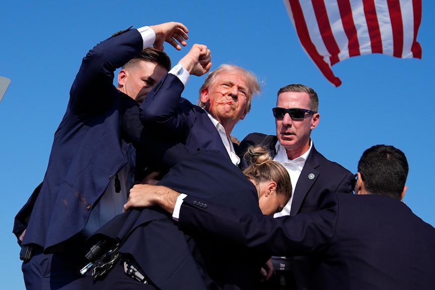 Donald Trump is surrounded by US Secret Service agents on a stage with red around his mouth