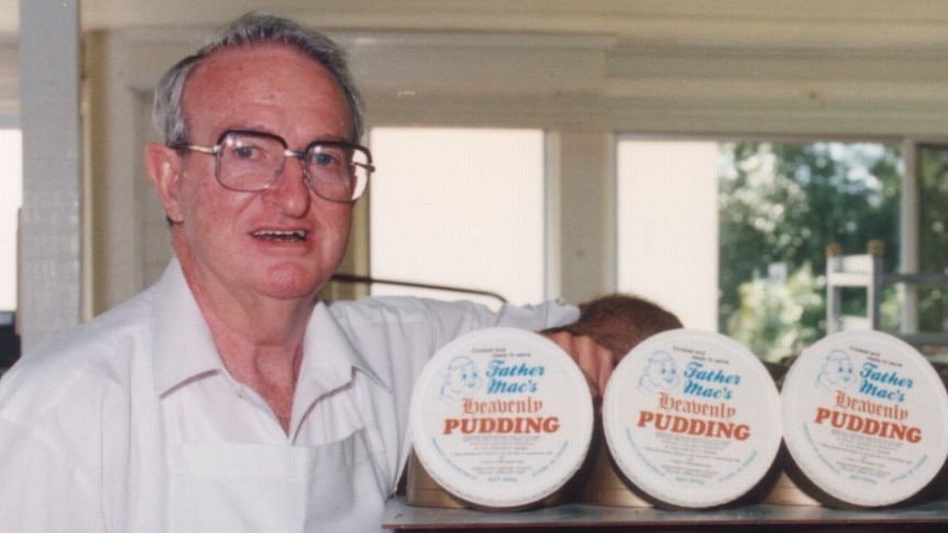 An older man with grey hair and a white shirt and glasses stands next to a stack of puddings in bronze containers.