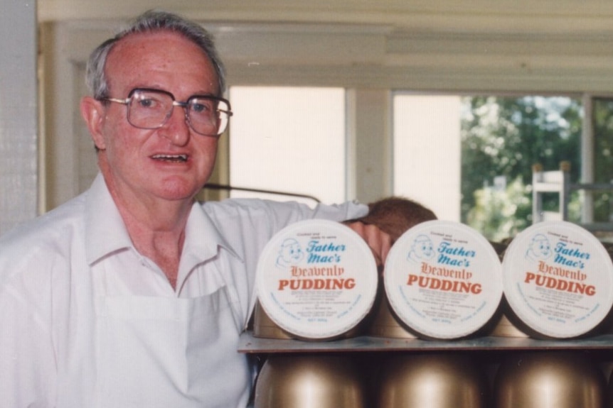 An older man with grey hair and a white shirt and glasses stands next to a stack of puddings in bronze containers.