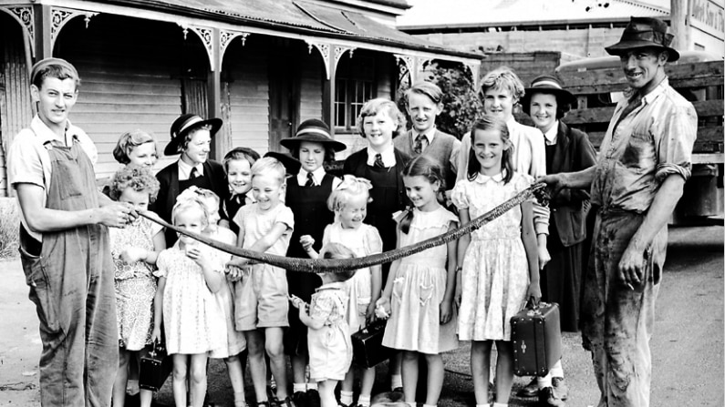 an old black and white photo of a group of children with men either side holding a very long black snake
