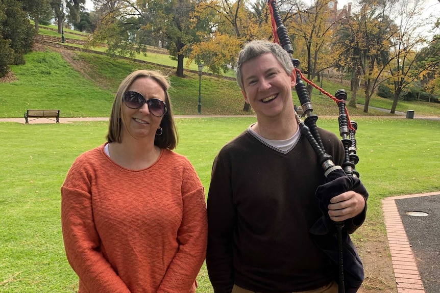 Kylie Webb and Brenton Hayward smile standing in a park. Brenton is holding a bagpipe.