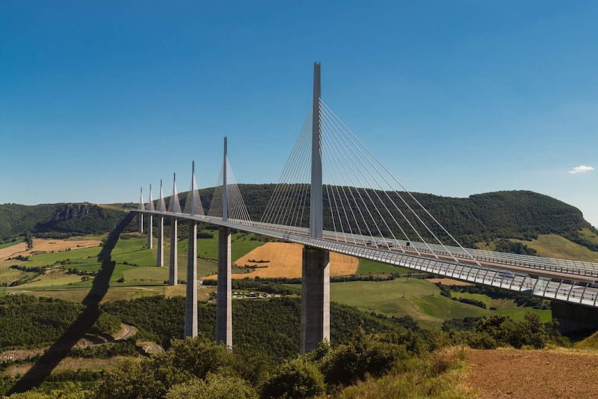 On a clear day, you view a white cable-stayed bridge over a hilly green landscape with its shadow drawing a curved line on it.
