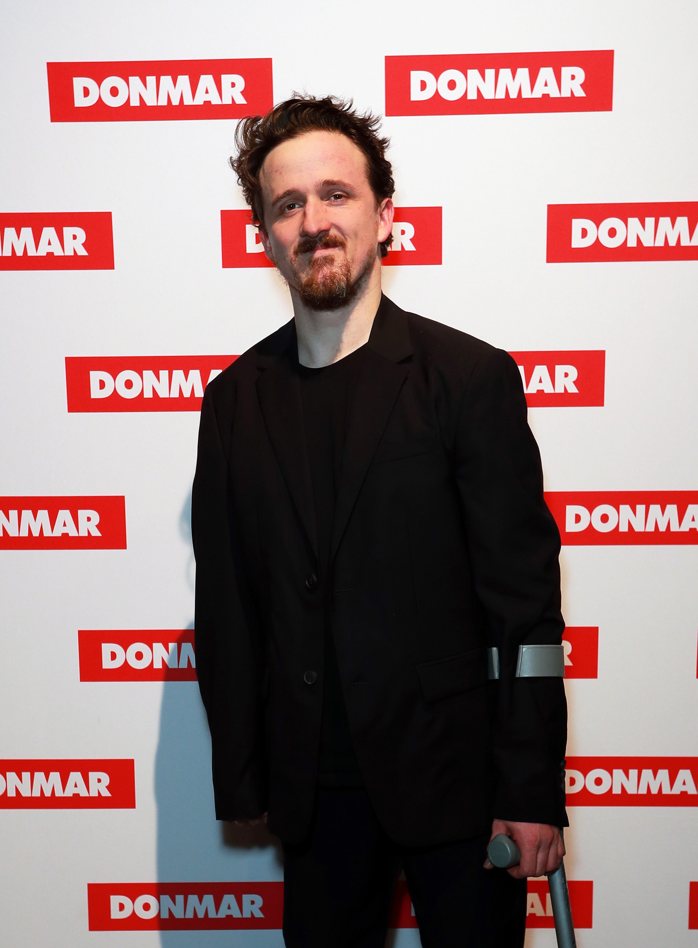 Daniel Monks, a caucasian man with dark brown hair and goatee, stands on a red carpet, he is using one crutch