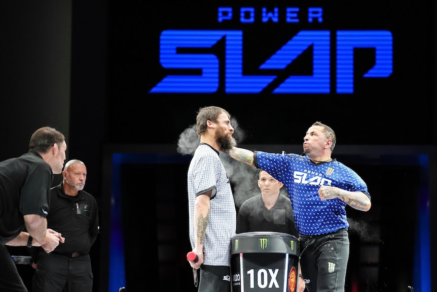 A man slaps another man as two referees look on. Behind them a big screen with the word 'slap' on it in blue.