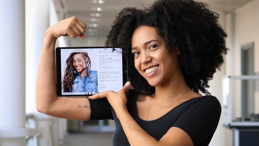 Young black woman smiles at camera while flexing her muscles and holding a photo of her sister.
