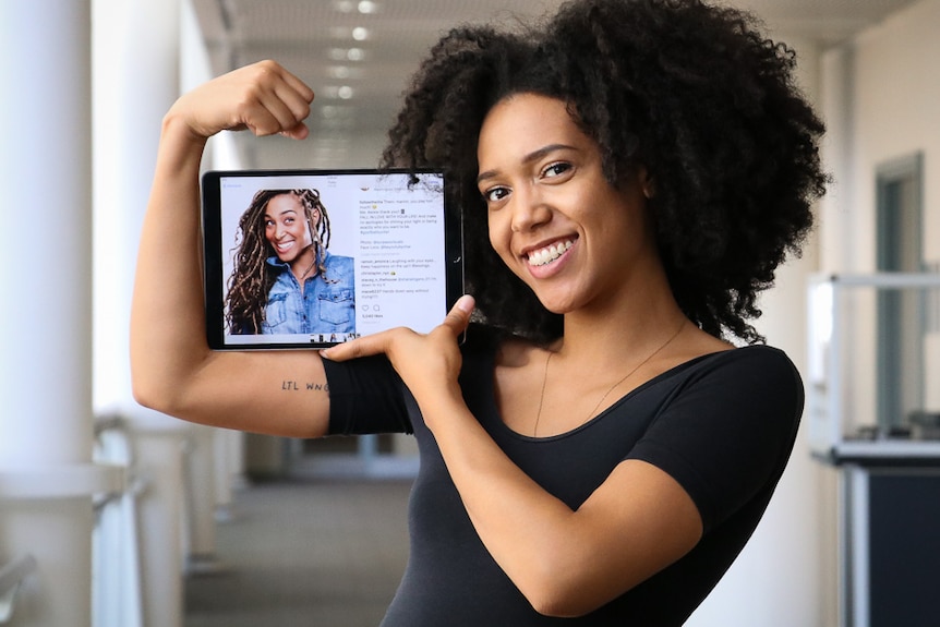 Young black woman smiles at camera while flexing her muscles and holding a photo of her sister.