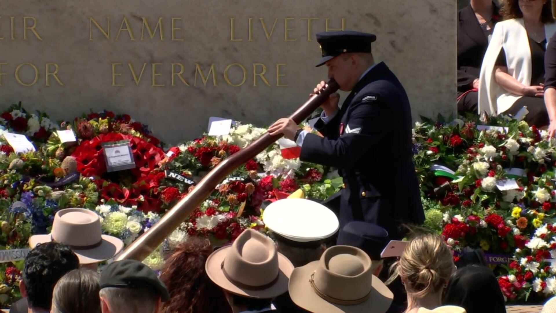 A man in military uniform plays didgeridoo in front of a military tribute stone.