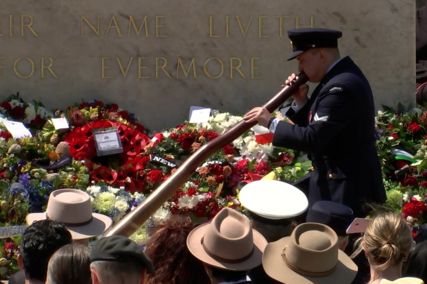 A man in military uniform plays didgeridoo in front of a military tribute stone.