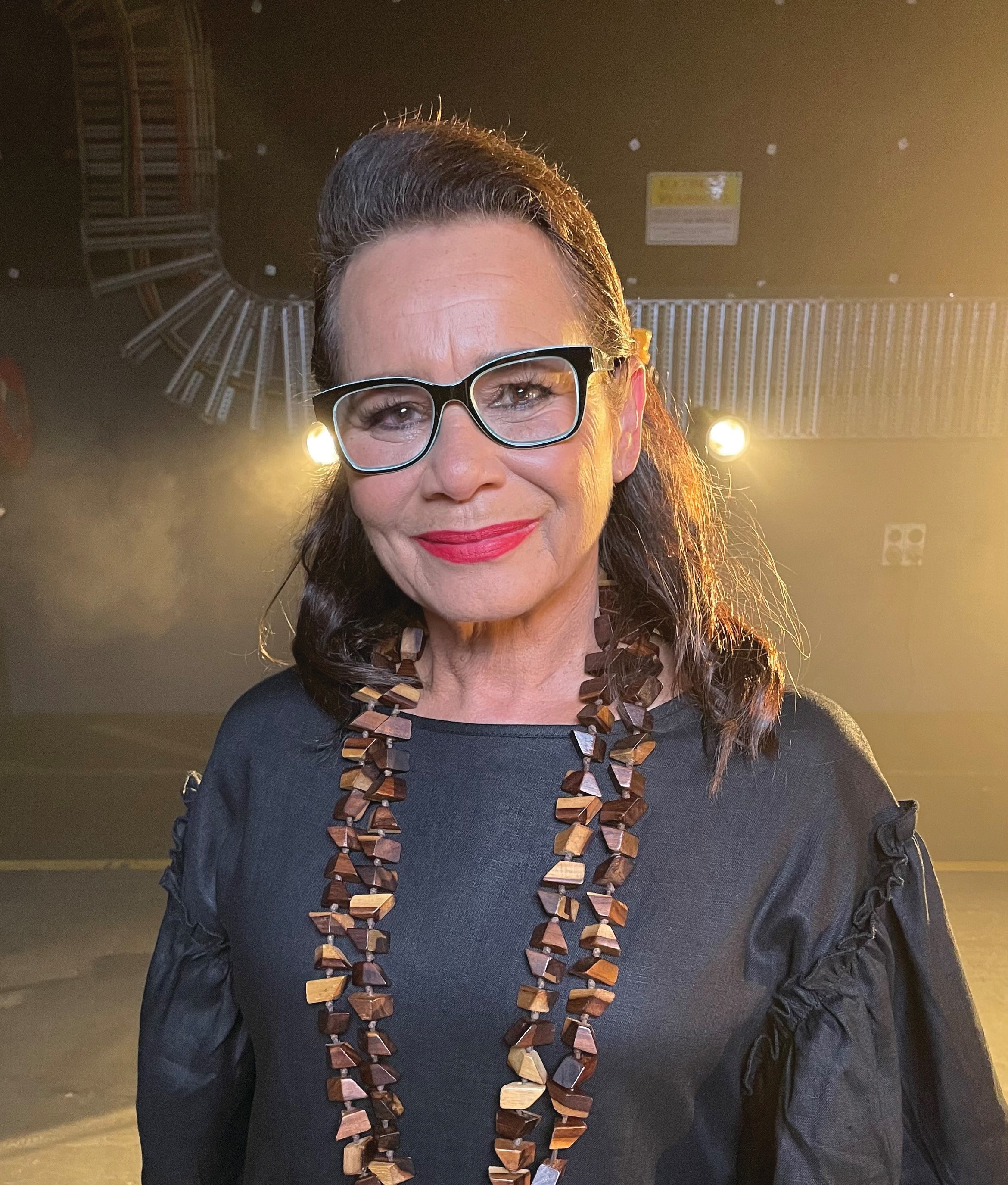 A portrait of Rhoda Roberts, she's wearing black glasses and a wooden necklace