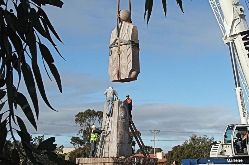 View through tree leaves of the top half of a stone person sculpture being connected to the basusing a crane with people helping