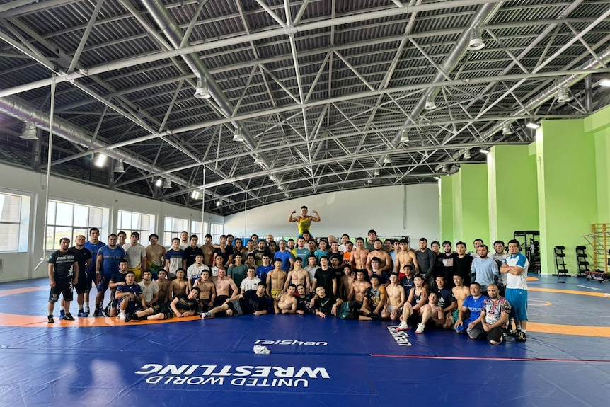 Big group of wrestlers in training smile for a photo in a large gym. There are approximately 80 people in the photo.