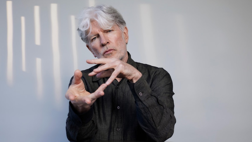 Tim Finn looks at the camera, his hands appear to cradle an invisible sphere