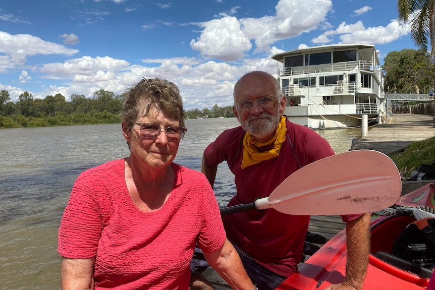 A man and a woman wearing pink shirts sit next to a kayak in front of a river. 