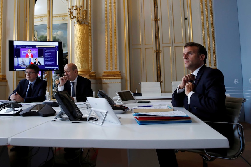Three men in suits sit in an ornate office and look attentively with a TV screen behind them.