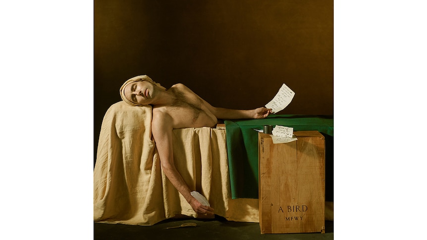 Andrew Bird lies naked in a casket with sheets of prose and a quill in his hands