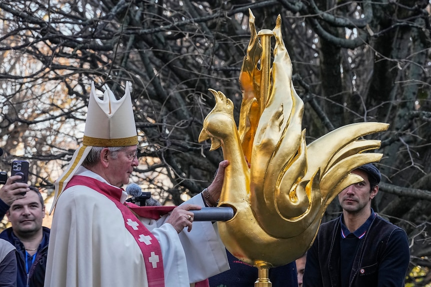 A man wearing a crown-like hat places a long pipe into a hole in a rooster sculpture.