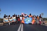 About 30 children walk in a line horizontally on the middle of the road with bright banners and blue cloudless sky behind them