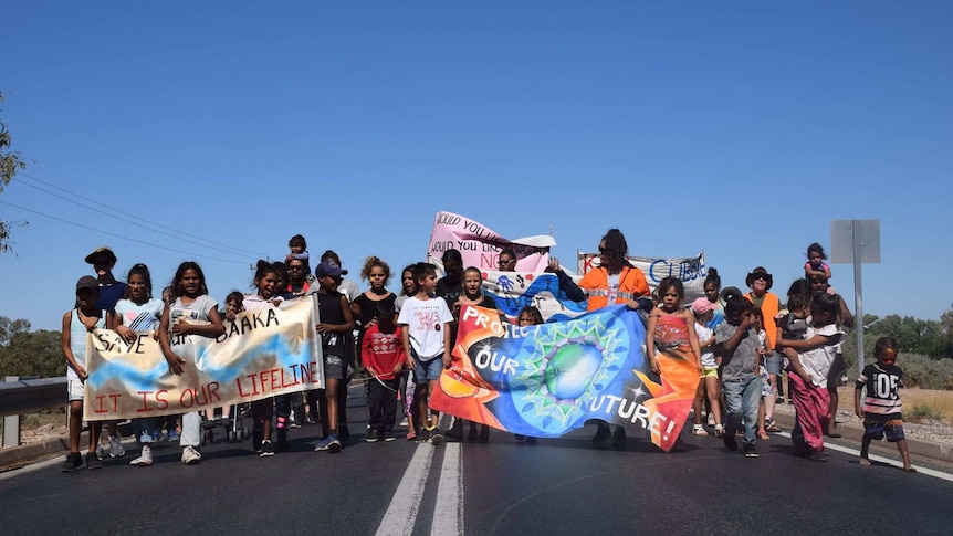 About 30 children walk in a line horizontally on the middle of the road with bright banners and blue cloudless sky behind them