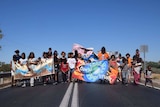 About 30 children walk in a line horizontally on the middle of the road with bright banners and blue cloudless sky behind them
