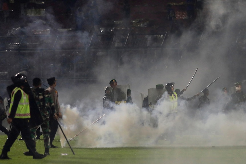 Police officers and soldiers stand on a football pitch amid tear gas smoke.