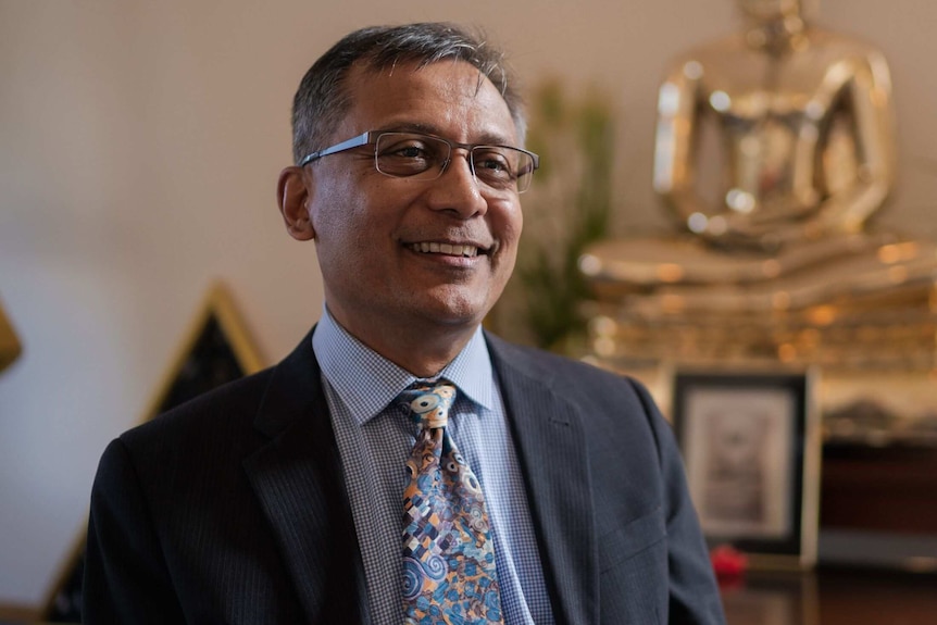 A man with glasses and a tie sitting in front of a gold buddhist statue.