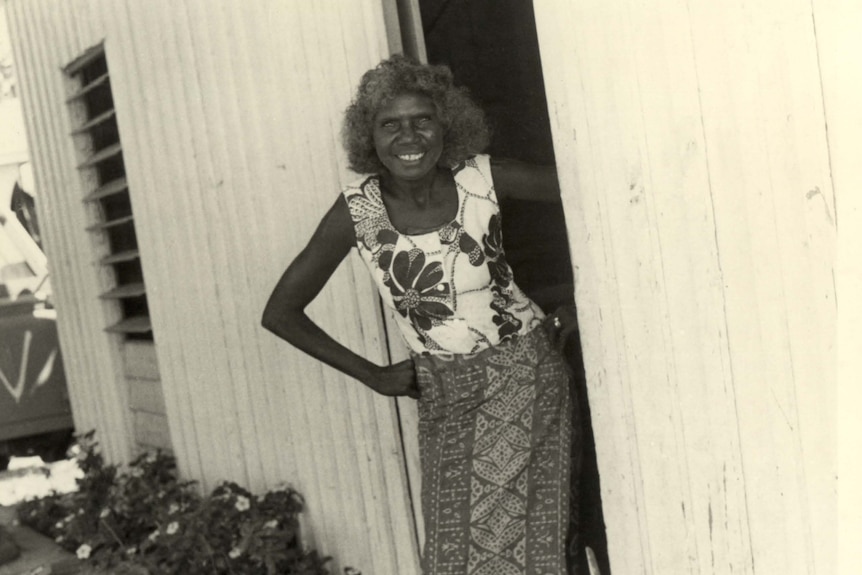 A black and white photograph of a woman standing at a doorway and posing. She places her hands on her hips and is smiling