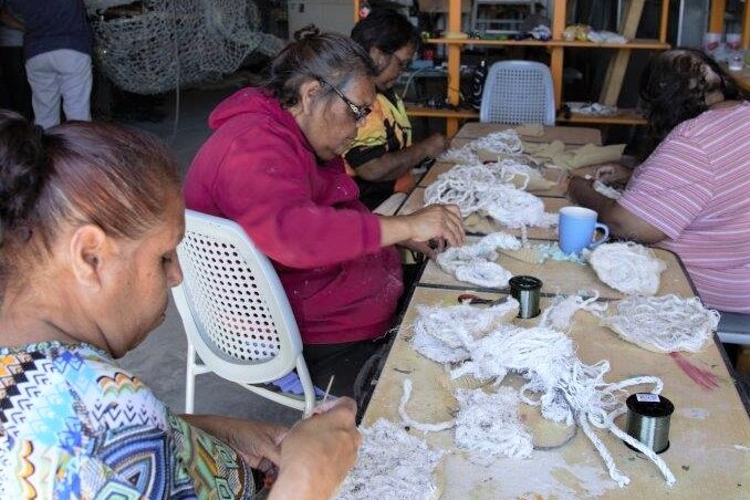 First nations women sitting at table sewing rope pieces, whale frame in the background.