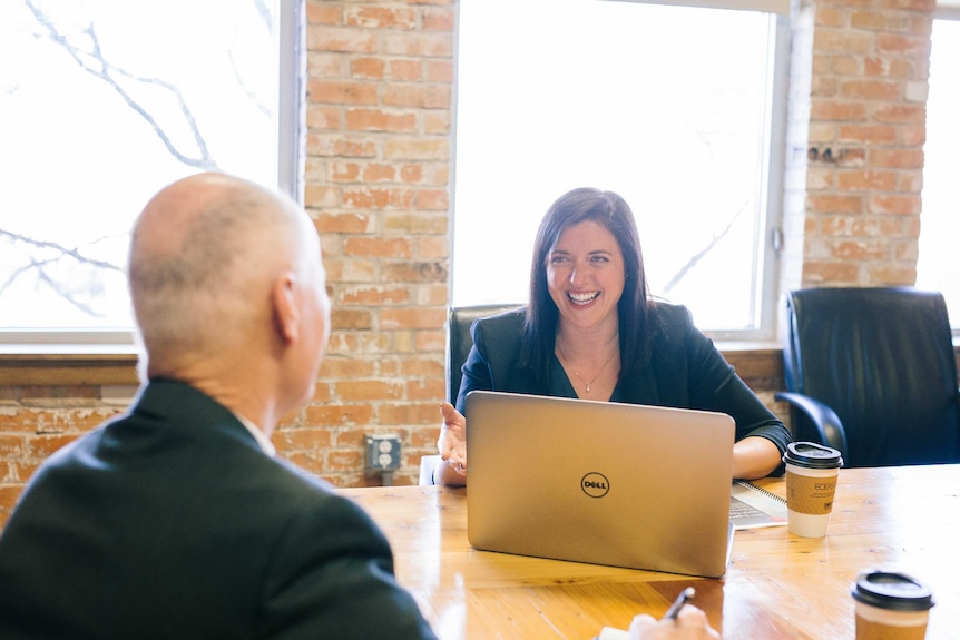 A woman sits in front of a laptop, smiling at a man.